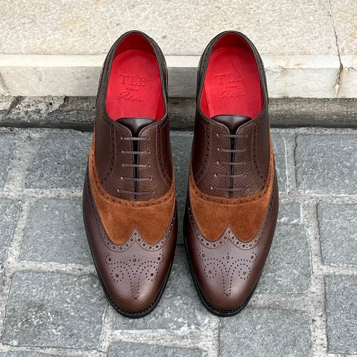 A pair of men's wingtip oxford shoes in dark brown leather and suede, featuring a red interior lining with the brand name 'TDS France' visible. The shoes are photographed from a top-down perspective on a gray stone paved surface with a light stone wall in the background.