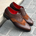 A pair of men's wingtip oxford shoes in dark brown and medium brown, featuring a textured leather upper, suede wingtip overlays, and a bright red interior lining, displayed on a gray stone pavement next to a concrete curb.