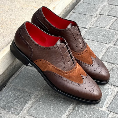 A pair of men's wingtip oxford shoes in dark brown and medium brown, featuring a textured leather upper, suede wingtip overlays, and a bright red interior lining, displayed on a gray stone pavement next to a concrete curb.
