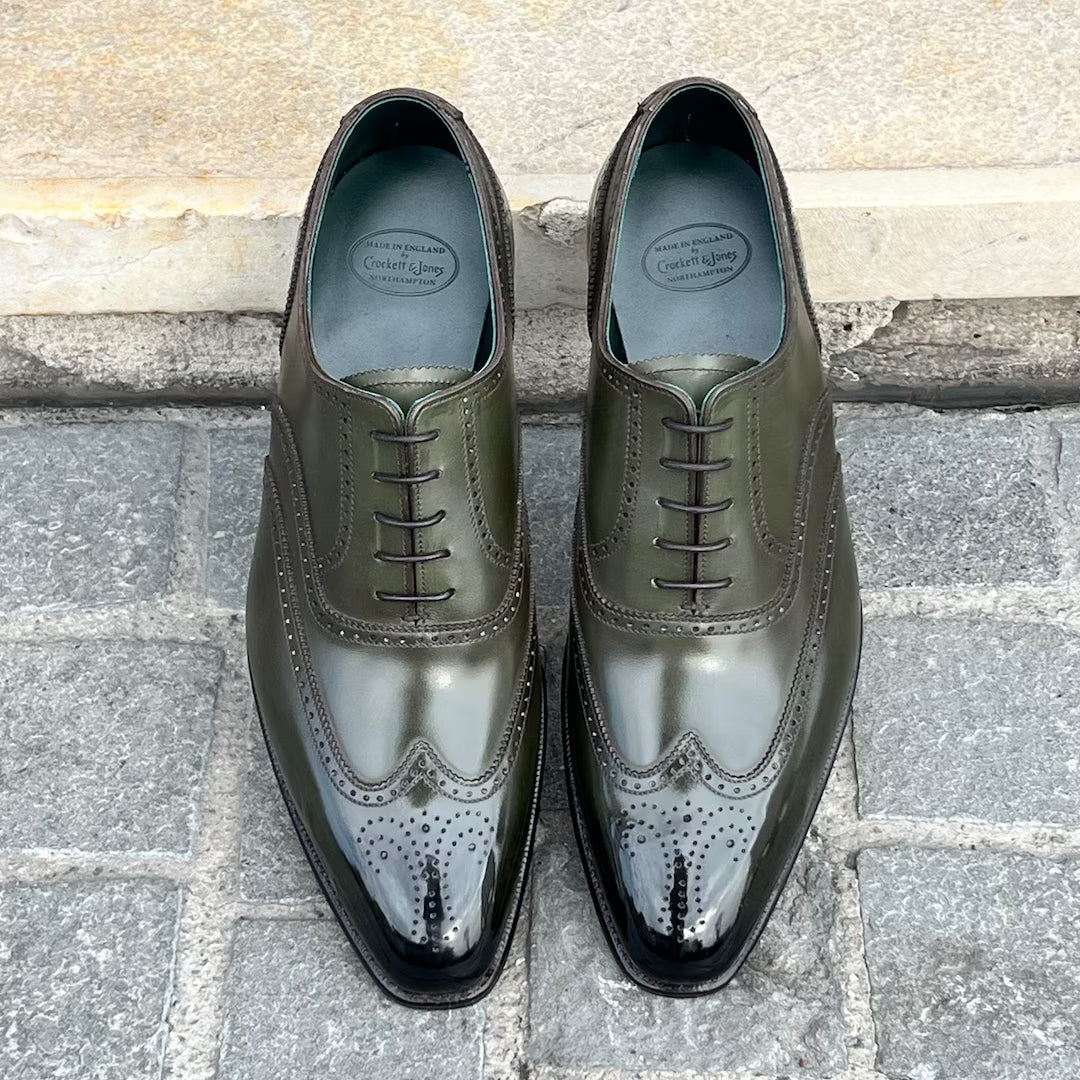 A pair of dark olive green wingtip oxford shoes with brogue detailing, displayed on a gray stone pavement against a light stone wall.