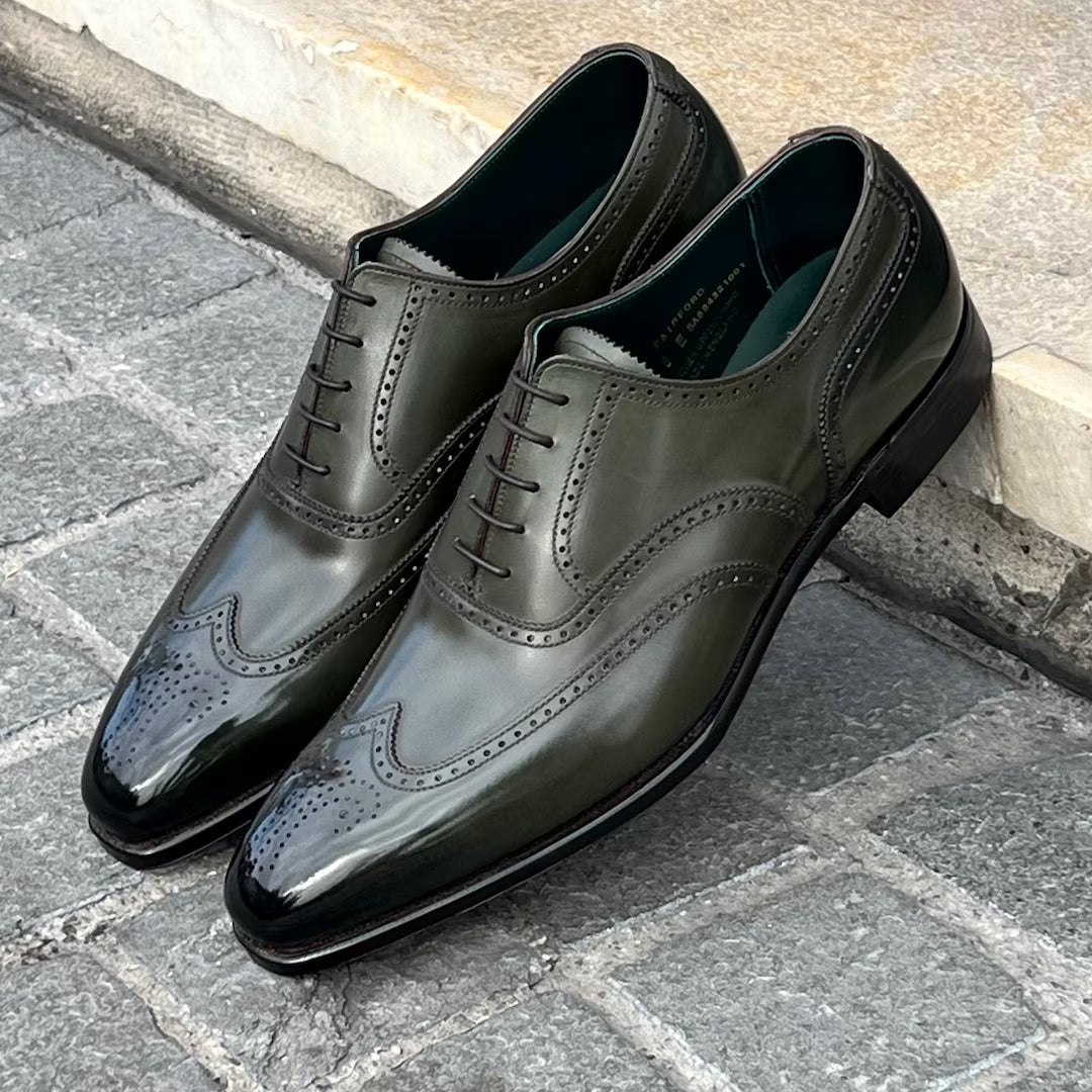 A pair of men's dark olive green leather wingtip oxford shoes with brogue detailing, displayed on a gray stone pavement next to a light-colored curb.