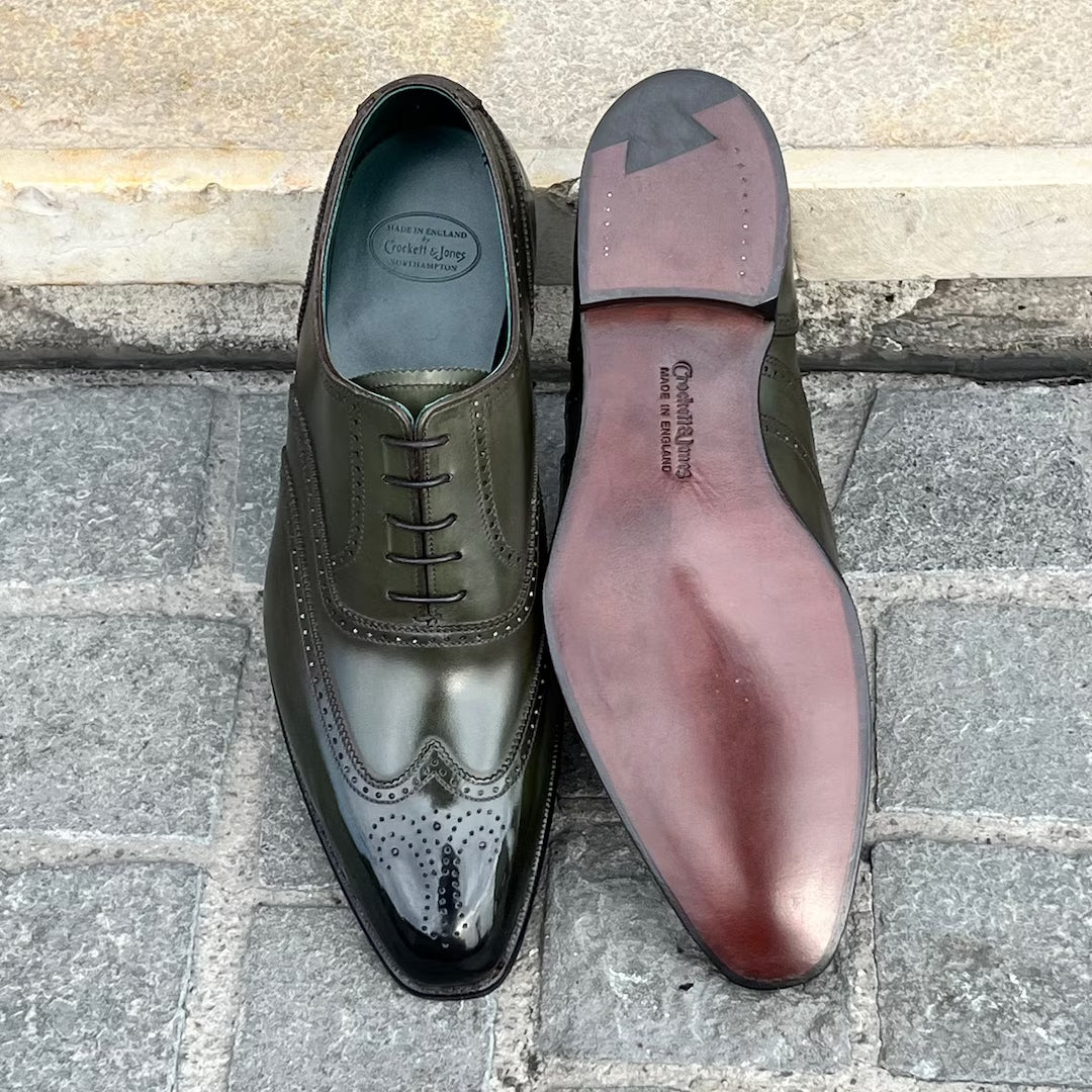 A pair of dark olive green wingtip oxford shoes shown on a stone pavement surface, with one shoe facing up to show the glossy brogue toe and the other showing the reddish-brown insole.