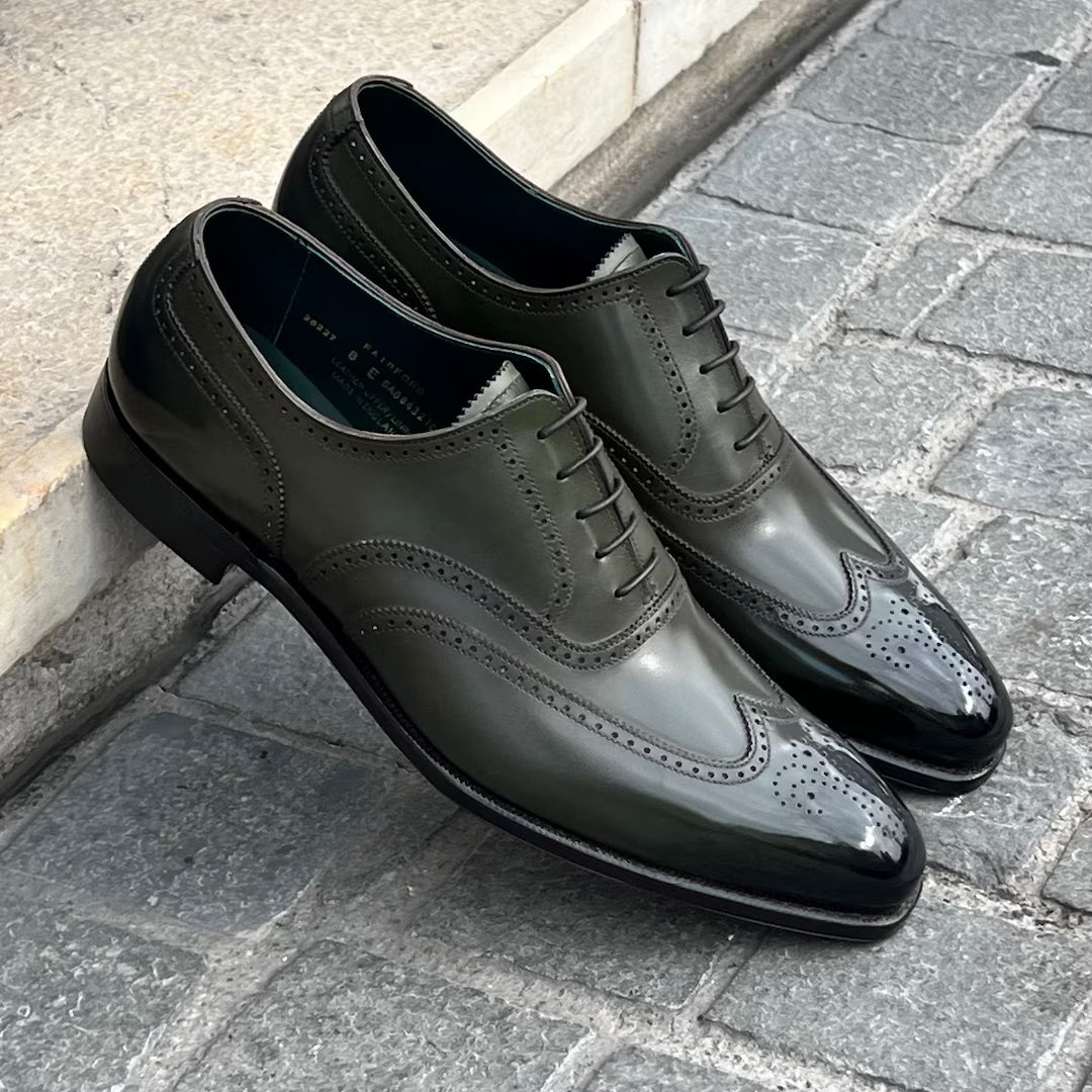 A pair of dark olive green glossy wingtip oxford shoes with perforated detailing, displayed on a gray stone pavement next to a light-colored curb.